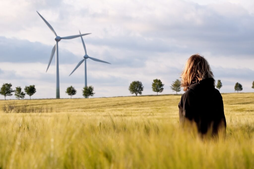 Eine Frau steht mit dem rücken zur Kamera in einem Weizenfeld und blickt auf zwei Windräder die am Horizont stehen.