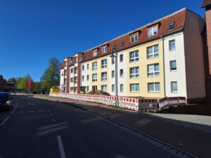 Eine Straße an der ein Mehrfamilienhaus steht. Vor dem Haus sind Baustellenbarken aufgebaut.