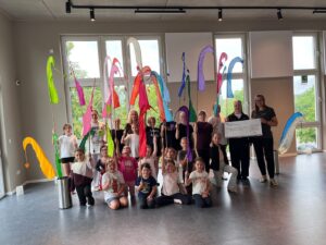 Eine Gruppe von Kindern steht in einem Saal und hält bunte Stoffe an Stöcken in die Höhe. Daneben stehen 2 Erwachsene die einen großen symbolischen Scheck übergeben.