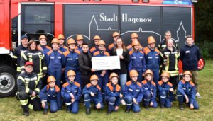 Mehrere Personen der Jugendfeuerwehr stehen in Montur und in drei Reihen vor einem Feuerwehrfahrzeug und nehmen einen symbolischen Scheck entgegen.