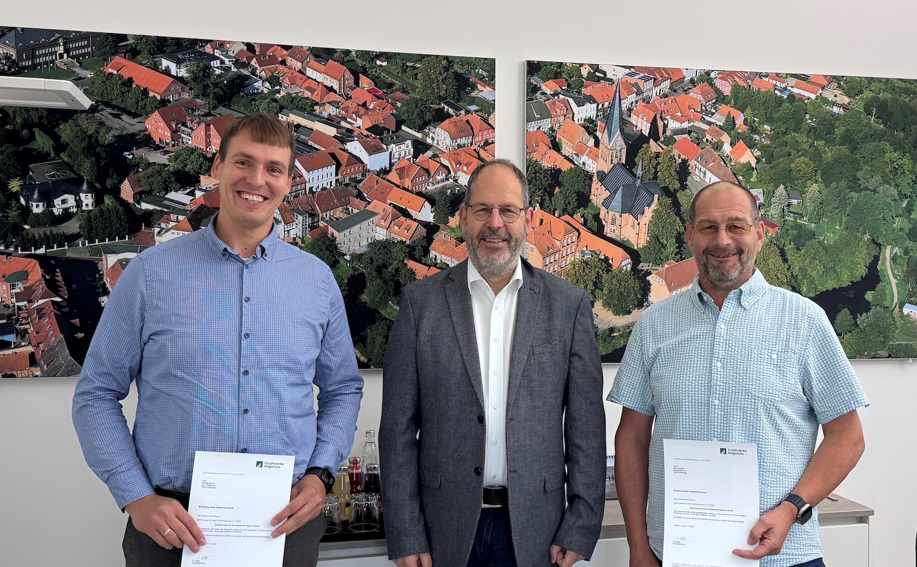 Drei Männer stehen in einem Büro vor einem großen Bild. Zwei der Männer halten eine Urkunde vor dem Körper.