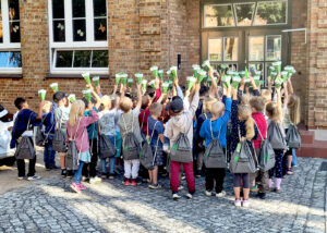 Eine Gruppe von Grundschulkindern steht mit dem Rücken zur Kamera vor einem Backsteingebäude und hält gemeinsam kleine, grüne Schultüten in die Luft.